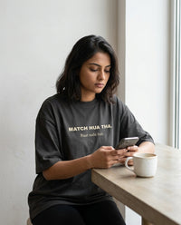 Indian woman sitting by a window scrolling her phone
with coffee wearing Match Hua Tha Baat Nahi Hui oversized
washed black tee — lifestyle shot | Aavitra