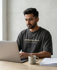 Indian man working on laptop with coffee and papers wearing 
Appraisal Mila Tax Ke Baad Kuch Nahi oversized washed black 
tee — lifestyle shot | Aavitra