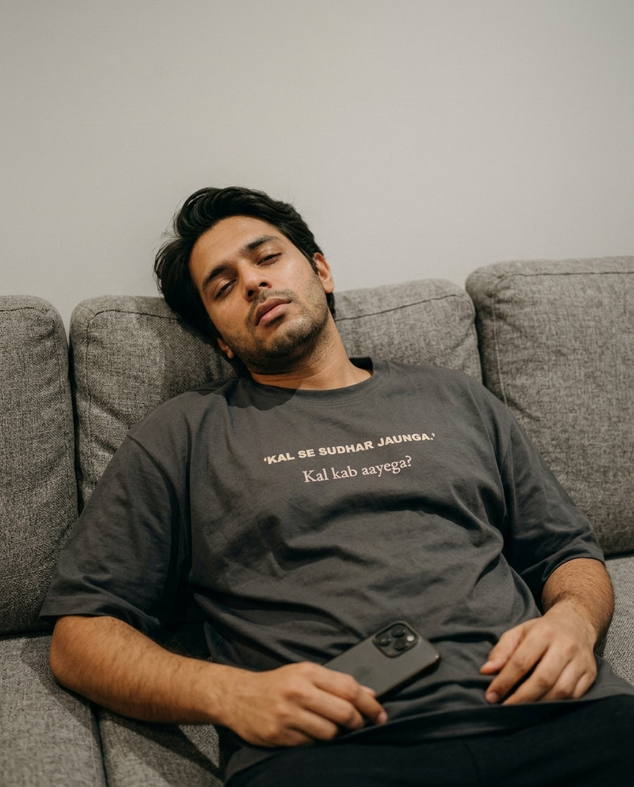 Indian man lying on a grey couch wearing Kal Se Sudhar 
Jaunga Kal Kab Aayega oversized washed black tee — 
lifestyle shot | Aavitra