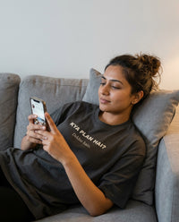 Indian woman relaxing on a grey couch scrolling her phone 
wearing Kya Plan Hai Dekhte Hain oversized washed black 
tee — lifestyle shot | Aavitra