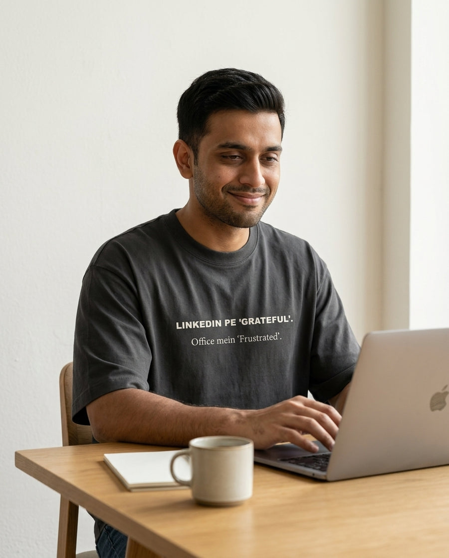 Indian man smiling at laptop with coffee wearing LinkedIn 
Pe Grateful Office Mein Frustrated oversized washed black 
tee — lifestyle shot | Aavitra