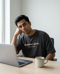 Indian man sitting at desk with laptop and coffee looking 
exhausted wearing Monday Aaya Main Nahi oversized washed 
black tee — lifestyle shot | Aavitra