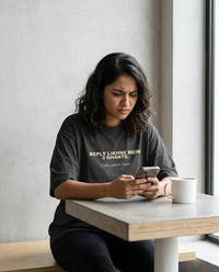 Indian woman frowning at her phone with coffee wearing 
Reply Likhne Mein 3 Ghante Unka Jawab Seen oversized 
washed black tee — lifestyle shot | Aavitra
